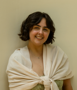 A person with short, dark, wavy hair smiles at the camera, wearing a light shawl over a checkered top, standing against a plain beige wall.