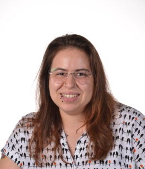 Person with long brown hair, glasses, and a patterned shirt smiles at the camera against a plain white background. A tattoo is visible on their left forearm.