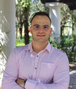 A man in a light pink striped shirt stands outdoors with arms crossed, smiling slightly, in front of white columns and greenery.