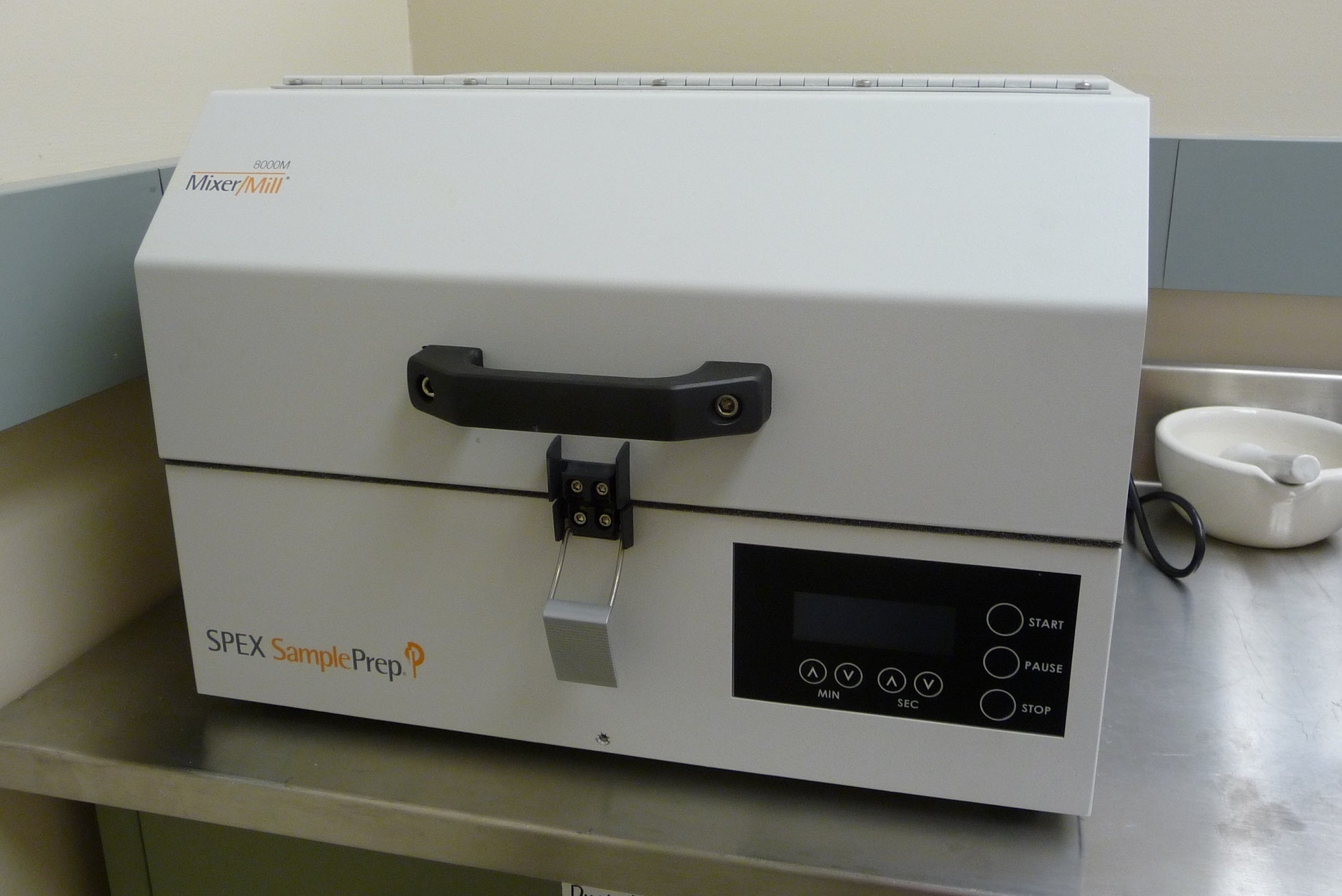 A SPEX SamplePrep Mixer/Mill 8000M laboratory machine on a stainless steel counter with a small white mortar and pestle in the background. The machine has a digital interface and several buttons.