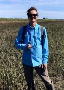 A person wearing sunglasses, a blue long-sleeve shirt, and a backpack stands in a grassy marsh, giving a thumbs-up gesture, with a car visible in the background.
