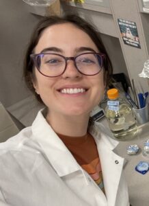 A person wearing glasses and a lab coat smiles in a laboratory setting, with labeled bottles and lab equipment visible on the counter behind them.