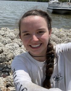A person in a white long-sleeve shirt takes a selfie near a pile of nets or ropes by the water, with a boat and trees in the background.