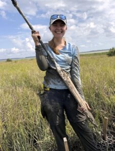 A person wearing outdoor gear, covered in mud, stands in a grassy marsh holding a long, muddy soil corer, smiling with tools clipped to their belt.