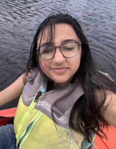 A person wearing glasses and a life jacket sits in a kayak on the water with their eyes closed and a slight smile.