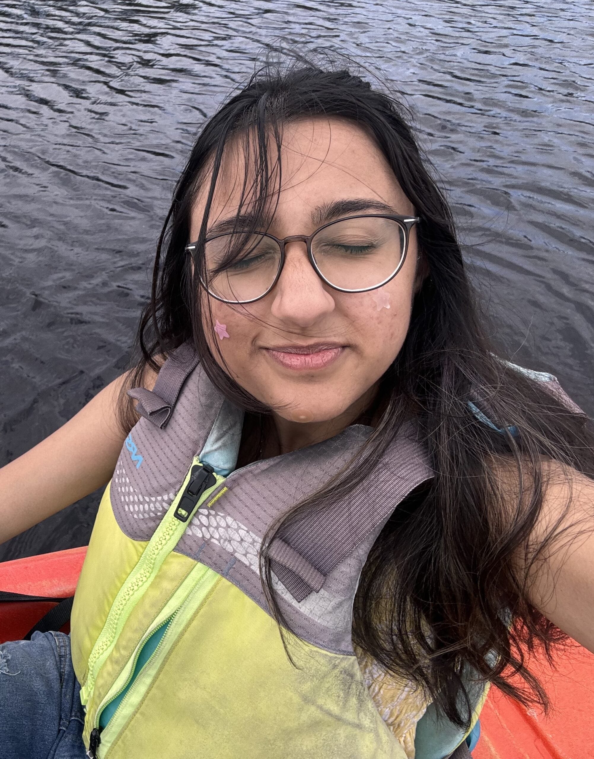 A person wearing glasses and a life jacket sits in a kayak on the water with their eyes closed and a slight smile.