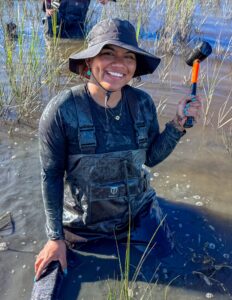 A person wearing waders and a hat kneels in muddy water, smiling while holding a mallet, surrounded by wetland grasses.