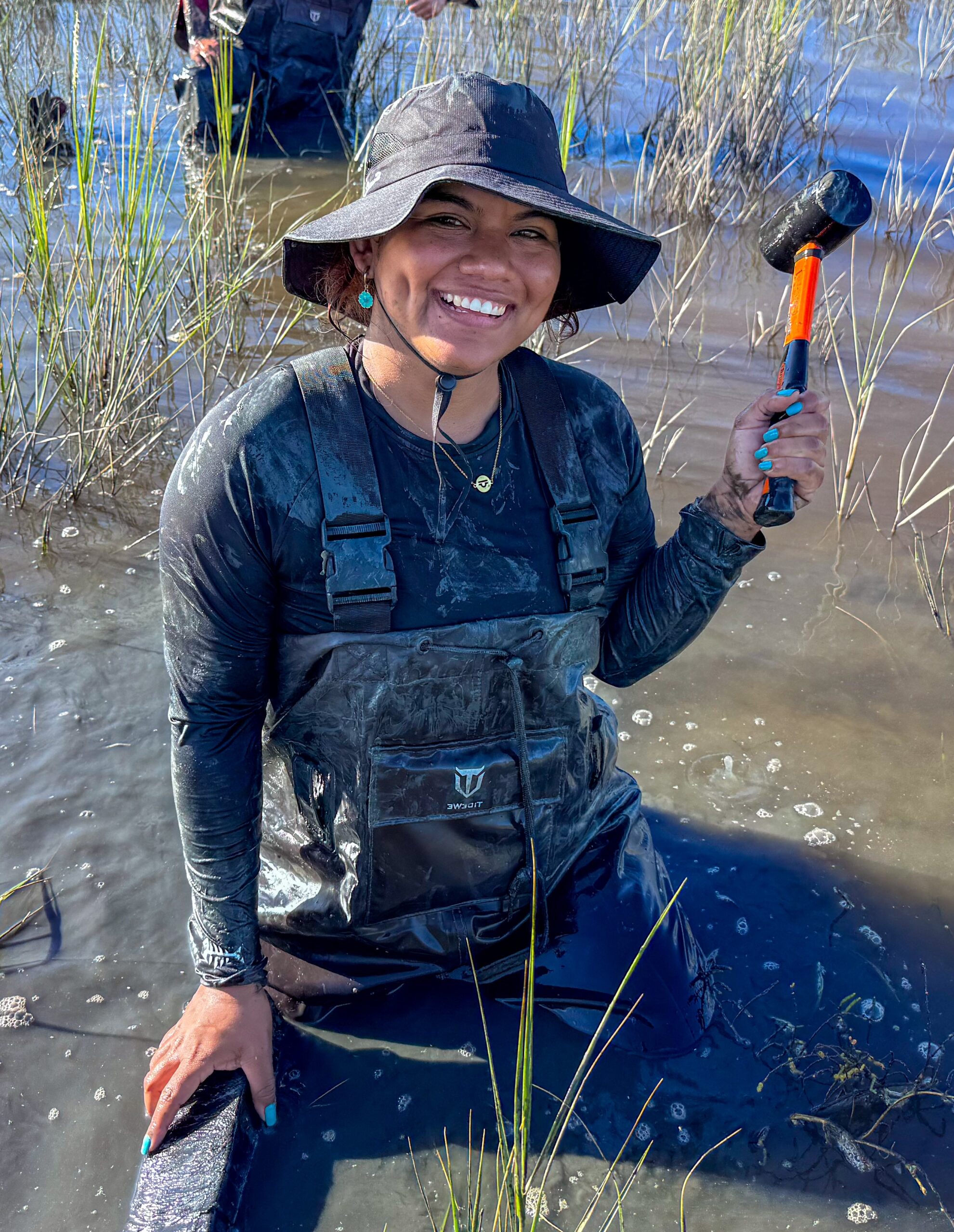 A person wearing waders and a hat kneels in muddy water, smiling while holding a mallet, surrounded by wetland grasses.