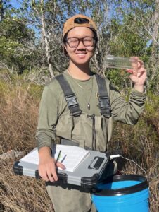 Person wearing glasses and outdoor gear holds a small clear container and a clipboard with a data sheet in a grassy natural environment.