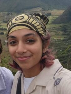 A person wearing a patterned headscarf smiles at the camera with green mountains and a cloudy sky in the background.