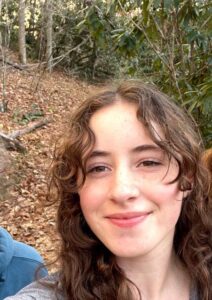 A young woman with wavy brown hair smiles at the camera while standing outdoors on a wooded trail with fallen leaves.