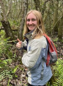 A person with long blonde hair, wearing a light jacket and red backpack, stands in a forest giving a thumbs up and smiling. Ferns and trees surround them.
