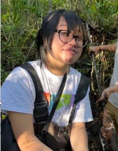 Person wearing glasses and a white T-shirt with a rainbow design, outdoors among green plants, with another person partly visible holding a stick.