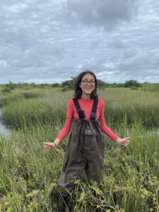 A person wearing glasses, a red long-sleeve shirt, and brown waders stands in tall grass near a marsh under a cloudy sky.