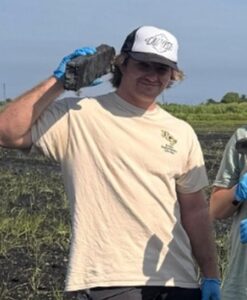 A person wearing a light t-shirt, black shorts, blue gloves, and a cap holds a dark rectangular object on their shoulder while standing in a grassy field.