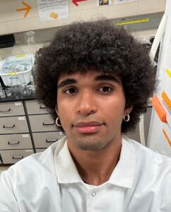 A person wearing a white lab coat takes a selfie in a laboratory with glassware, drawers, and scientific equipment visible in the background.