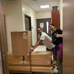 Two people are standing in a doorway, smiling, while one person pushes a cart loaded with large cardboard boxes.