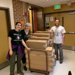 Three people smiling and standing next to a cart filled with stacked cardboard boxes in a hallway.