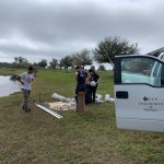 People standing near equipment by a pond and a white van labeled "UCF Department of Biology" on a grassy field under a cloudy sky.