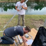 Two men assemble metal poles and other parts near a pond. One man stands holding a pole while the other kneels, referring to instructions. A black bag lies nearby.