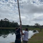 Two individuals are seen assembling a tall, metallic structure on a grassy area near a lake, under a partly cloudy sky.