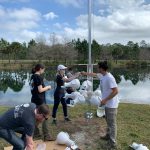 A group of people adjust equipment on a tall pole near a pond, setting up what appears to be a research or monitoring device in an outdoor environment.