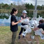 Two people are assembling and attaching white equipment to a metal pole near a pond, with another person assisting nearby.