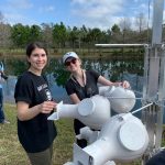 Two people stand outdoors by a body of water, both smiling and operating white scientific equipment. The background features trees, grass, and a partly cloudy sky.