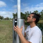 A person in a white t-shirt is adjusting a tall metallic structure outdoors with a grassy field and trees in the background.