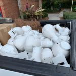 A pickup truck bed filled with numerous white plastic streetlight covers and several cardboard boxes, parked near a brick building and some greenery.