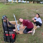 Three people sit outdoors on cardboard assembling equipment. One person smiles while holding a component. They are on grass near a small body of water. Equipment and tools are visible around them.