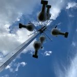 A tall pole against a blue sky with white clouds, featuring multiple birdhouses attached by rods near the top.