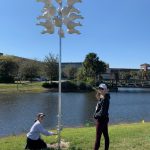 Two people adjusting a pole-mounted weather instrument by a pond on a sunny day, with buildings and trees in the background.