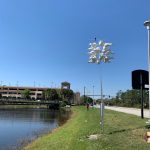 A cluster of birdhouses is mounted on a pole near a pond with a grassy shoreline. In the background, there is a bridge and a multi-story parking structure. The scene is set on a clear, sunny day.