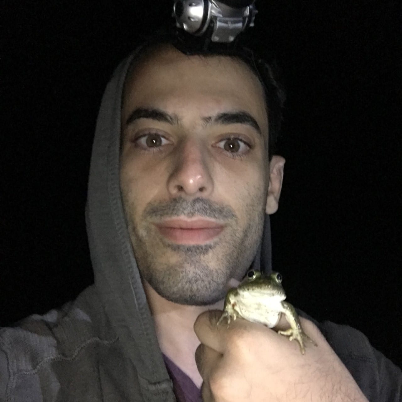 A person wearing a headlamp and a hoodie holds a frog in their hand, posing for the camera in a dark setting.