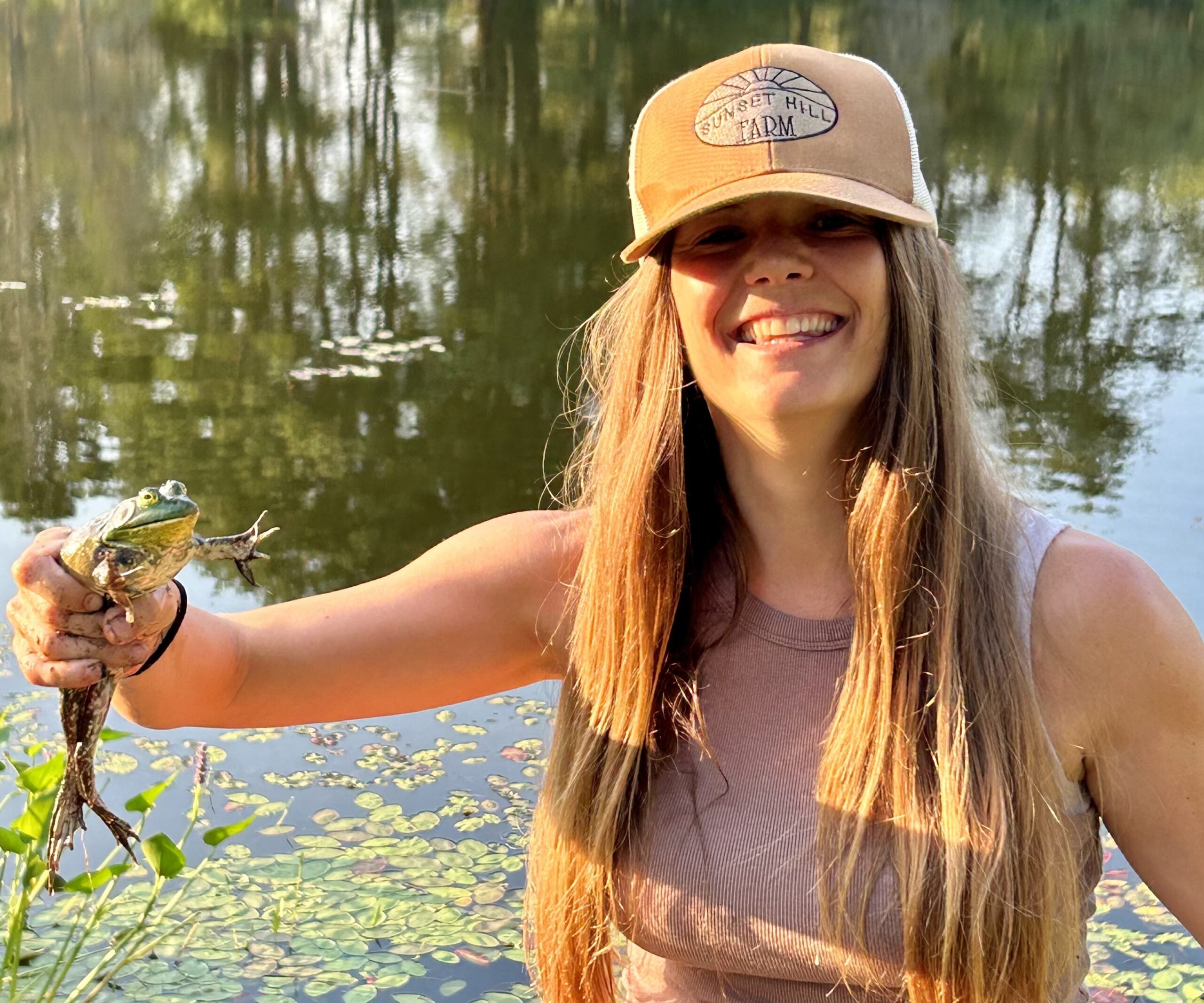 A woman stands by a pond, smiling and holding a frog. She wears a tan cap and a sleeveless top.