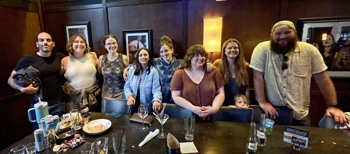 A group of nine people, including one child, are posing together in a restaurant. The table in front is cluttered with drinks and plates. They are smiling at the camera.