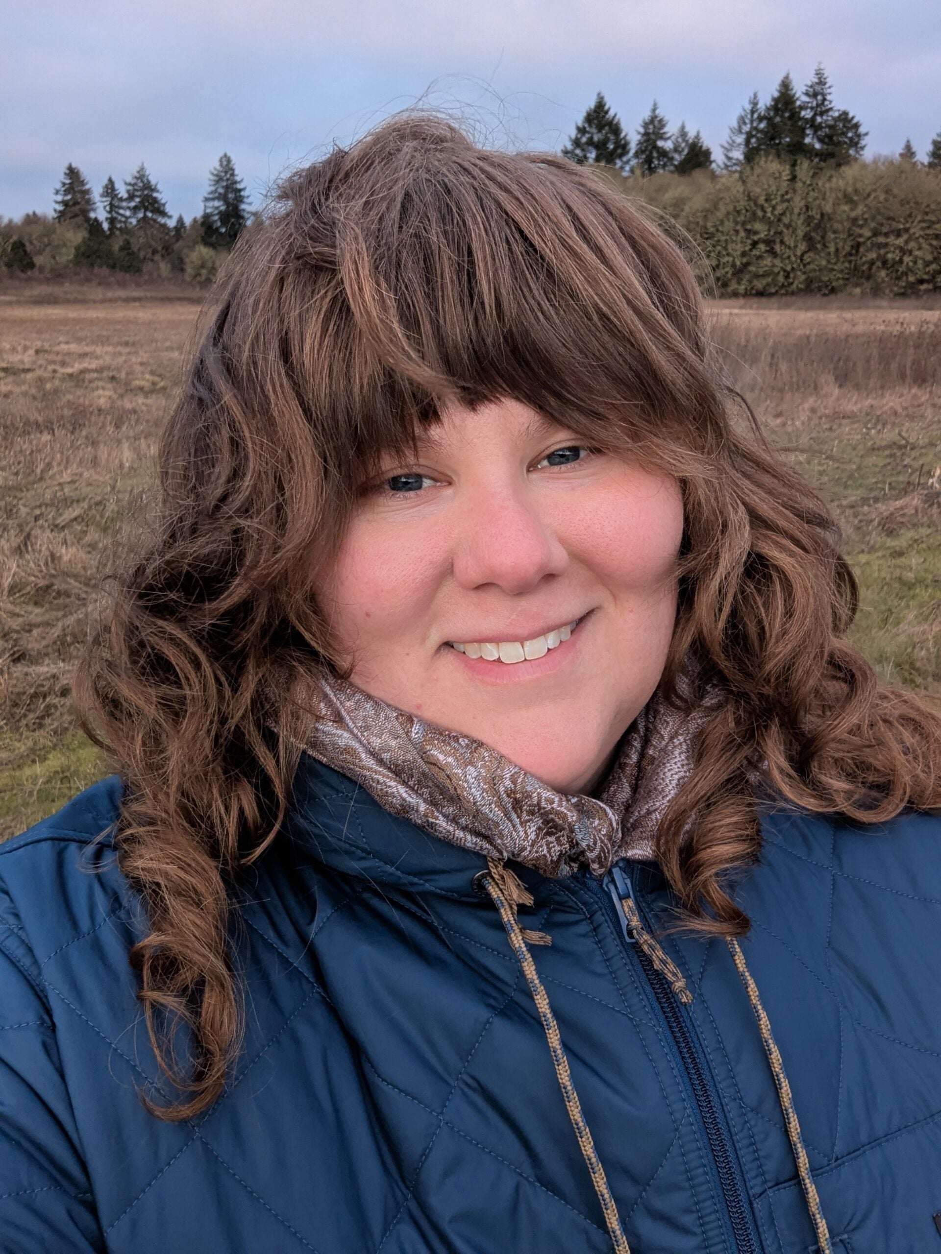 A person with wavy brown hair wearing a blue jacket and scarf smiles outdoors in a field with trees in the background.