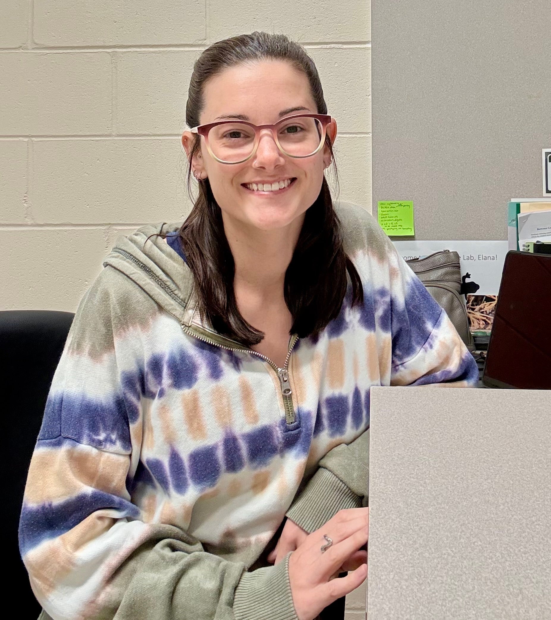 A woman with glasses and a tie-dye hoodie sits and smiles at a desk with office supplies and papers in the background.