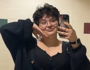 Person with short curly hair and glasses poses for a mirror selfie in a tiled room, wearing a black sweater and holding a phone with a patterned case.