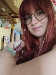 A person with long red hair and glasses holds a green praying mantis on their arm indoors.