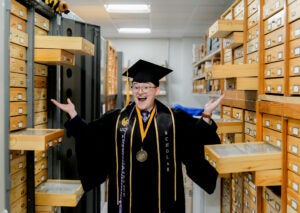 A graduate in cap and gown stands smiling with arms raised in a room filled with open wooden specimen drawers.