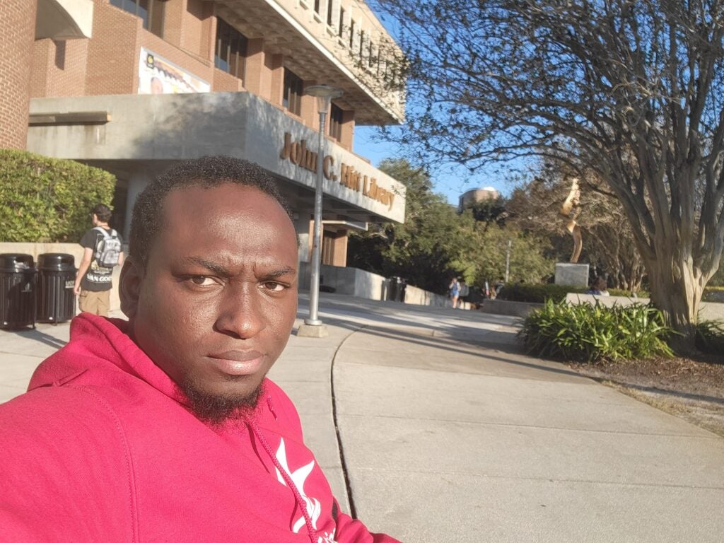 A man in a red hoodie sits outside a building labeled “John C. Hitt Library” on a sunny day.