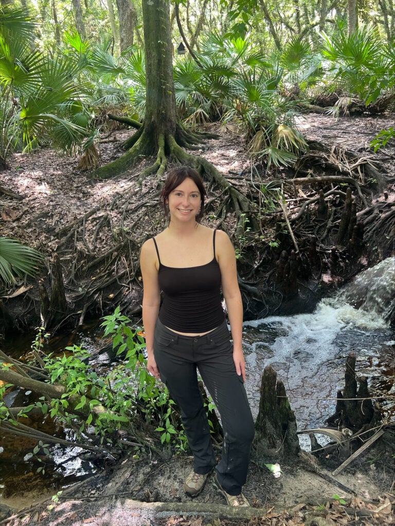 A woman stands by a small stream in a forest, wearing a black tank top and dark pants. Lush green plants and trees surround her, and sunlight filters through the leaves.