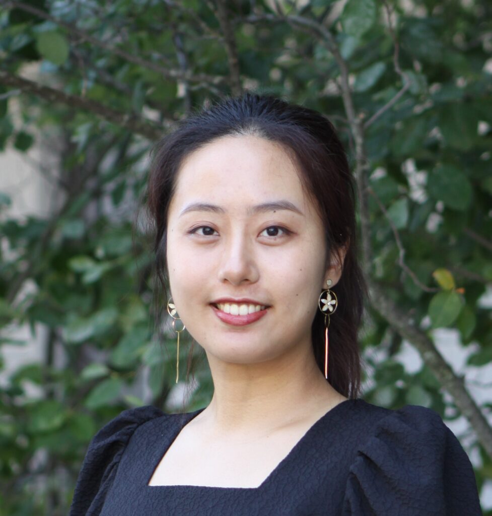 Woman with long dark hair wearing a black dress and dangling earrings, smiling in front of leafy green background.