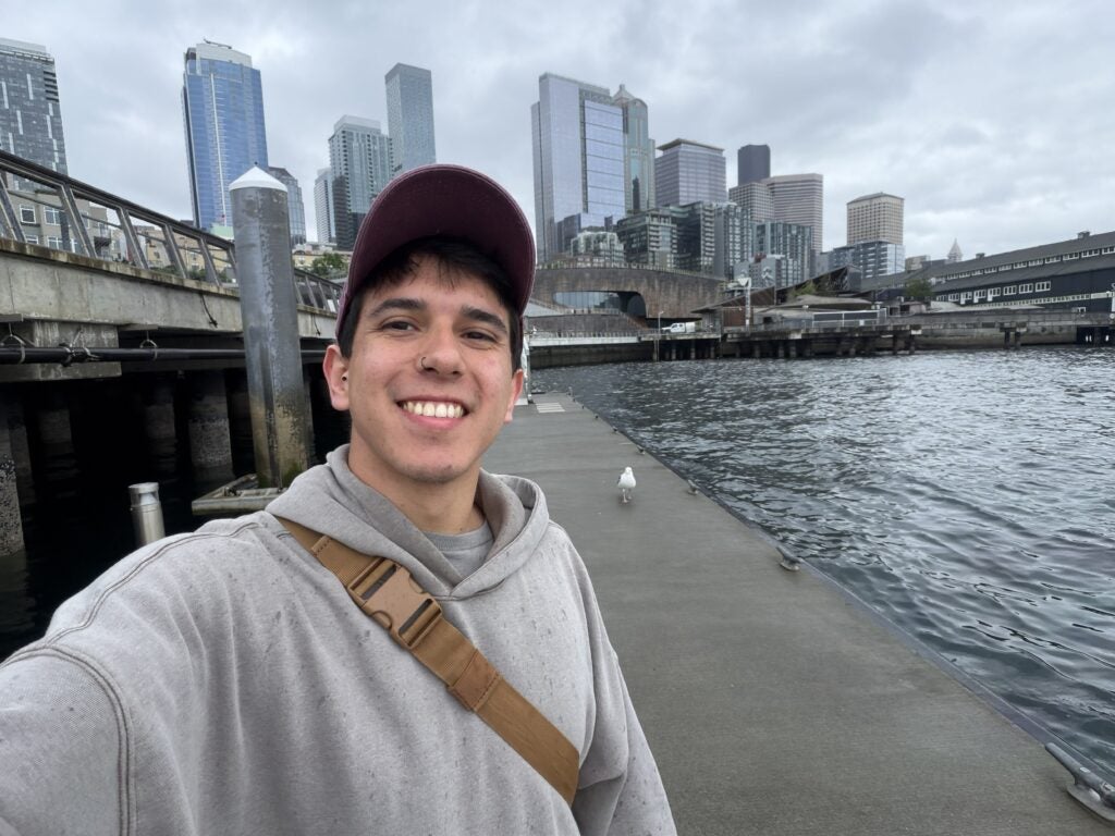 A person in a hoodie and cap takes a selfie on a waterfront dock with city skyscrapers in the background under a cloudy sky.