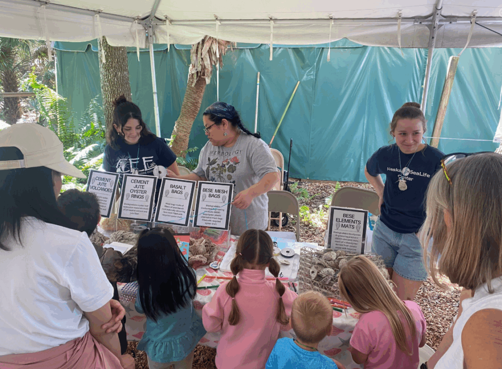 A group of children and adults gather around a table under a tent, engaging with marine science-communication-outreach displays and interactive materials presented by three women.