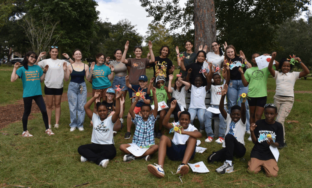 A group of children and adults poses outdoors in a park during a science-communication-outreach event, holding up crafts and drawings, smiling at the camera with trees and grass in the background.