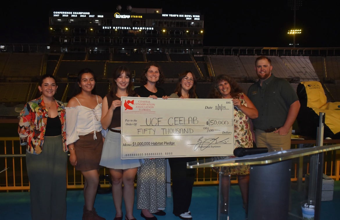 A group of seven people stand at night in a stadium, holding a large $50,000 check made out to UCF CEELAB from Coastal Conservation Association Florida for undergraduate-graduate-research initiatives.
