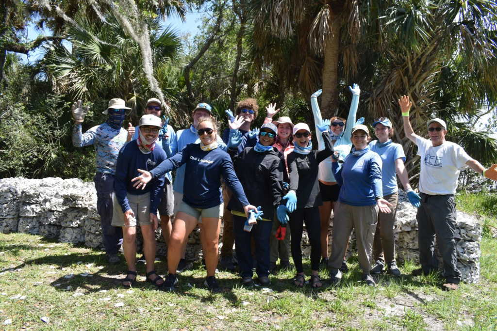 A group of people involved in undergraduate-graduate-research, outdoors and wearing hats, sunglasses, and gloves, stand together smiling and waving in front of palm trees and a pile of restoration materials